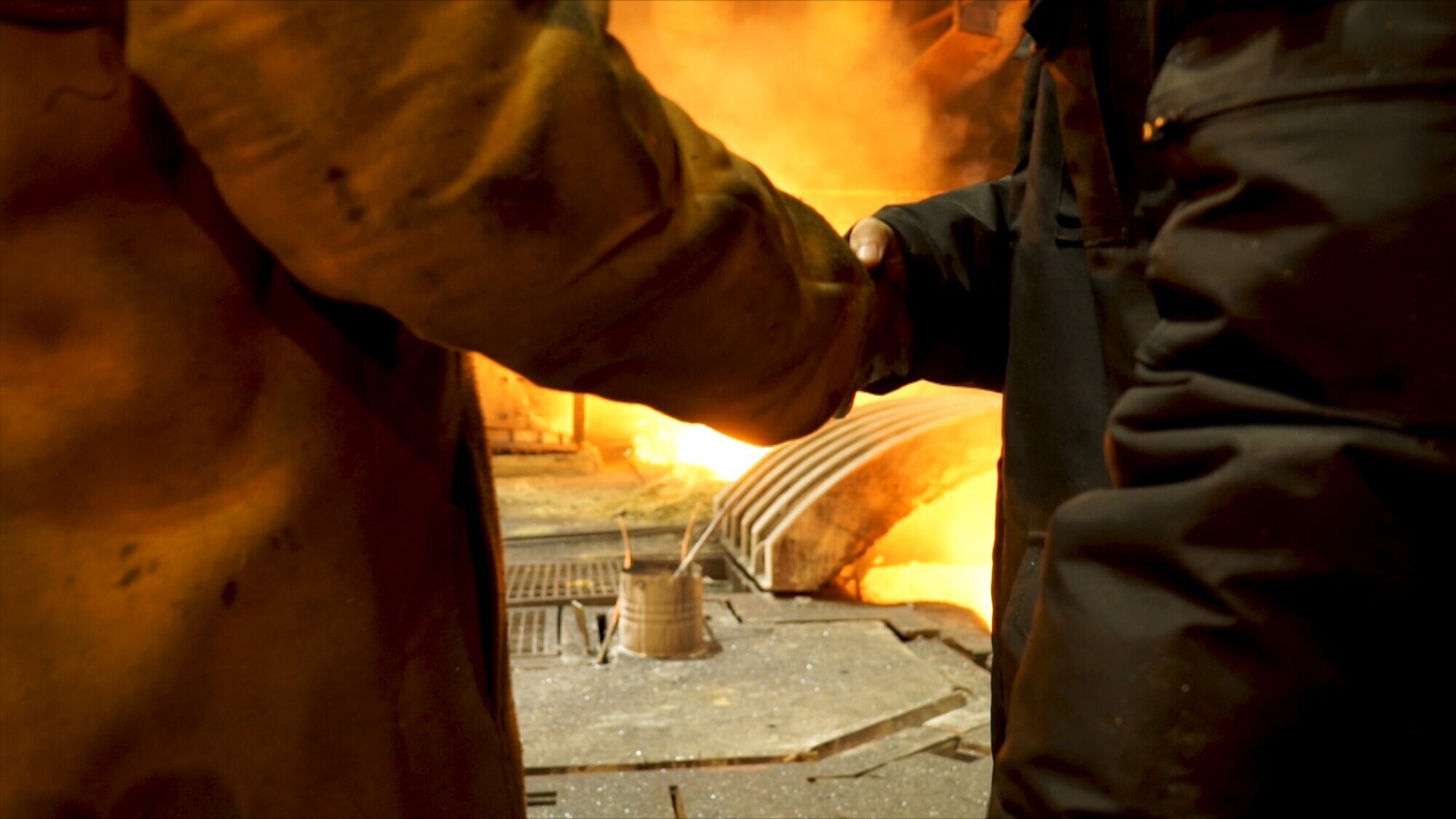 Rear view of two steelmakers at ingot casting shaking hands in front of electric arc furnace in hot shop, metallurgical production. Stock footage. Heavy industry and agreement concept.